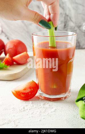 Un verre transparent avec du jus de tomate rouge et une main de femme tient une branche de céleri. Concept d'alimentation saine. Orientation verticale. Sélectif FO Banque D'Images