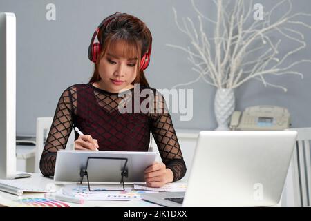 Portrait à la taille du joli graphiste asiatique assis au bureau et en écoutant de la musique dans un casque tout en travaillant avec un numériseur Banque D'Images