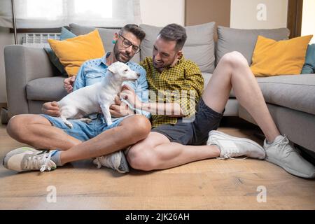 Couple gay heureux assis sur le sol et jouant avec le chien à la maison. Photographie de haute qualité Banque D'Images