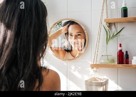 Belle femme hispanique regardant dans le miroir dans la salle de bains écologique blanche. Shleves en bois et bouteilles de cosmétiques réutilisables. Concept de wellnes Banque D'Images