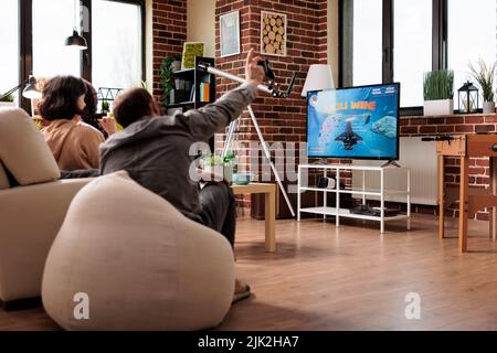 Des personnes multiculturelles assises sur un canapé dans la salle de séjour tout en jouant à des jeux de simulation d'espace sur la console. Divers amis assis à la maison tout en appréciant des jeux vidéo ensemble. Banque D'Images