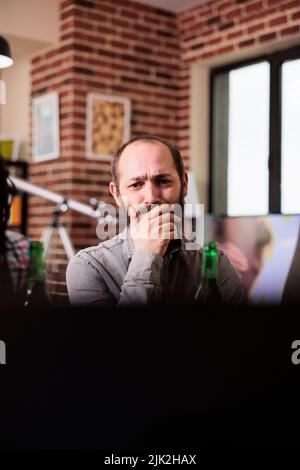 Inquiet caucasien homme assis à la maison tout en regardant les événements tragiques de l'actualité vidéo en ligne sur ordinateur portable. Personne inquiète assise dans le salon tout en regardant des films de suspense sur un ordinateur portable moderne. Banque D'Images
