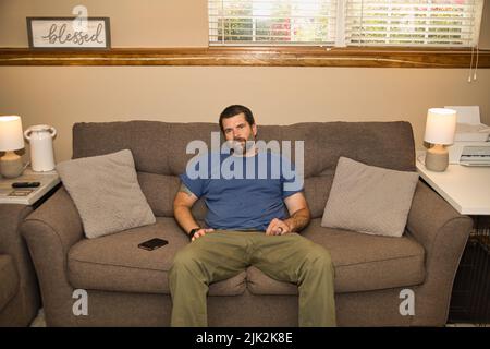 Homme barbu en chemise bleue se détendant sur un canapé gris. C'est tout simplement froid. Banque D'Images