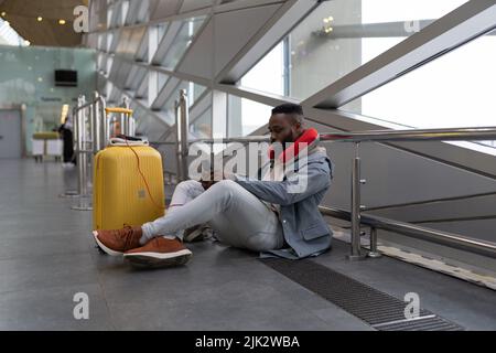 Un jeune homme noir sérieux est assis à pieds croisés sur le sol en attendant de prendre l'avion en utilisant la banque électrique pour téléphone portable Banque D'Images