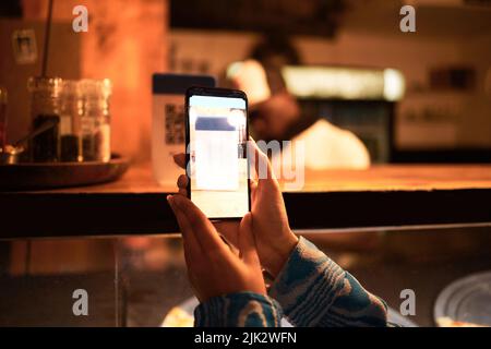 Personne avec appareil photo de téléphone scannant code-barres QR pour le menu en ligne dans un restaurant, magasin ou café moderne la nuit. Main d'une femme qui prend une photo de numérisation pour Banque D'Images