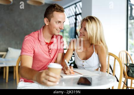 Des étudiants heureux assis dans la table de café de l'université et faisant des devoirs ou de la rédaction de plans. Une jeune femme et un homme passent du temps ensemble pendant le frein du café, en prenant des notes. Banque D'Images