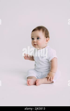 Vue latérale de bébé bébé enfant en couches et body blanc est rampant sur tous les quatre avec ses jambes dehors, essayant de se tenir debout regardant l'appareil photo au-dessus de wh Banque D'Images