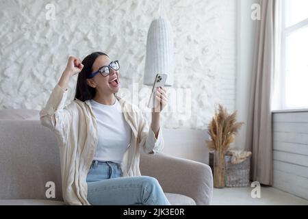 Une femme heureuse à la maison souriante et heureuse de recevoir un message, une brune assise sur un canapé portant des lunettes et tenant le téléphone à la main criant gaiement Banque D'Images