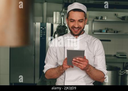 Chef souriant en uniforme faisant l'ordre dans la tablette numérique pour le fournisseur debout sur la cuisine Banque D'Images