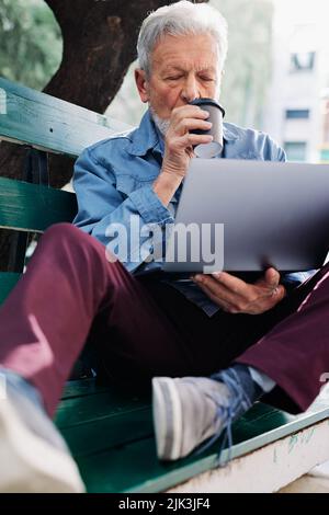 Un étudiant senior s'assoit sur le banc dans un parc, sirotant un café et tient un ordinateur portable sur ses genoux. Étudiants seniors en recyclage. Banque D'Images
