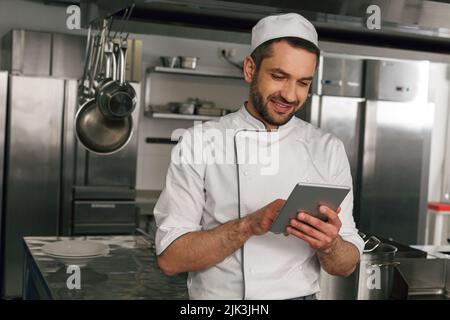 Chef souriant en uniforme faisant l'ordre dans la tablette numérique pour le fournisseur debout sur la cuisine Banque D'Images