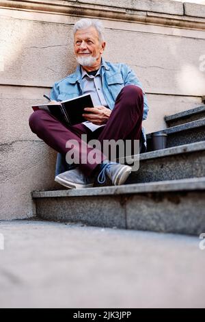 Un étudiant senior heureux s'assoit sur les escaliers devant le bâtiment de l'université et écrit les devoirs. Programme de recyclage pour les aînés. Banque D'Images