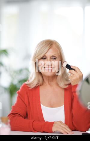 Une belle femme blonde senior est assise à la maison et fait son maquillage matin routine. Femme âgée mettant blush sur son visage Banque D'Images
