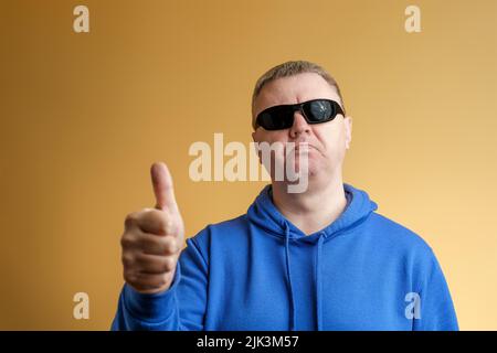 Un homme montre un geste de pouce vers le haut. Portrait d'un homme dans un sweat à capuche bleu et des lunettes de soleil Banque D'Images