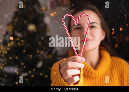 Femme souriante dans un chandail orange avec des cannes rouges et blanches en forme de coeur près de l'arbre de noël. Banque D'Images