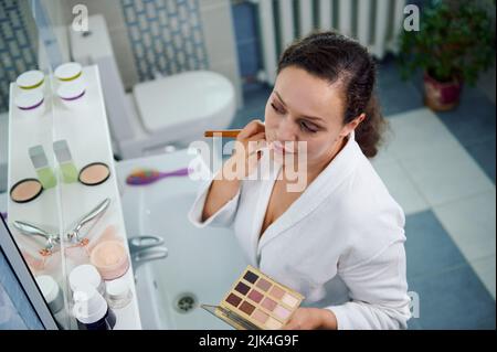 Femme étourdissante applique de la poudre matifiante sur son visage, satisfait de son apparence, regarde son reflet dans le miroir Banque D'Images