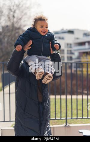 Joyeux et joyeux petit garçon afro-américain de 5 élevé par sa mère bien-aimée, tous deux portant des vêtements d'hiver et se tenant près de la clôture en métal - zone résidentielle de bâtiments avec arrière-plan de parc. Photo de haute qualité Banque D'Images