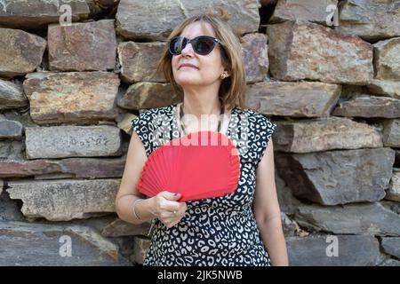 Portrait d'une femme âgée penchée sur un mur de pierre à l'extérieur et avec un ventilateur rouge en raison de la chaleur estivale. Banque D'Images