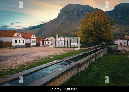 Vue majestueuse sur la rue rurale avec maisons traditionnelles en rangée et fontaine dans le centre du village. Village de Torocko et montagne Piatra Secuiului à Backgro Banque D'Images