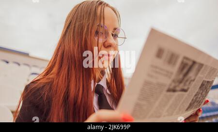 Une femme en lunettes et un costume de commerce lit un journal tout en étant assise au stade. Banque D'Images