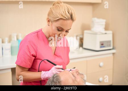 Femme souriante dermatologue chirurgien élimine le néoplasme à l'aide de scalpel à ondes radio Banque D'Images