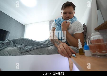 Un jeune homme atteint d'un mal de gorge se trouve dans le lit et atteint un thermomètre Banque D'Images