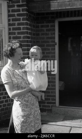 1960s, historique, à l'extérieur de l'entrée d'une maison, une mère portant une robe de l'époque, tenant sa fille de bébé, Angleterre, Royaume-Uni. Banque D'Images