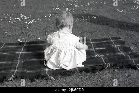 1960s, historique, printanier et extérieur, sur un terrain d'herbe couvert de pâquerettes, une vue de derrière un chlld bébé assis sur un tapis à motif tartan, Angleterre, Royaume-Uni. Banque D'Images