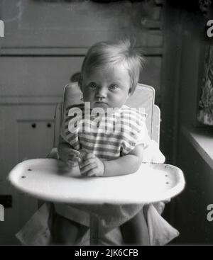 1960s, historique, enfant en bas âge assis dans une chaise haute, avec un plateau métallique en émail, tenant un biscuit, Angleterre, ROYAUME-UNI. Banque D'Images