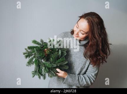 Adulte belle femme souriante quarante ans avec brunette cheveux bouclés en robe tricotée grise chaude avec couronne de noël en forme de sapin à la main sur le dos gris Banque D'Images