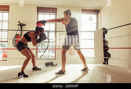 Boxeur féminin qui esquive une frappe de son entraîneur personnel dans un anneau de boxe. Jeune femme athlétique ayant une séance d'entraînement de boxe dans une salle de gym. Banque D'Images