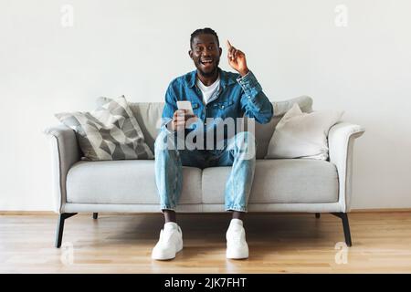 Un homme afro-américain enthousiaste tenant un smartphone ayant une idée assis à l'intérieur Banque D'Images