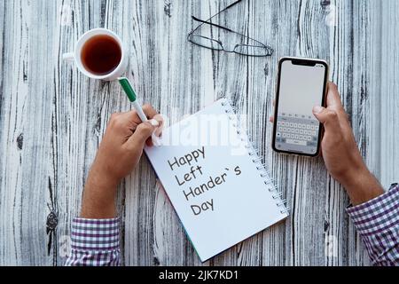 Homme écrit dans un bloc-notes. Happy Left hander écrit le texte du jour par sa main gauche dans un bloc-notes tenant une maquette d'écran de téléphone. Verre et tasse de thé. Photo conceptuelle du jour de la gauche Banque D'Images