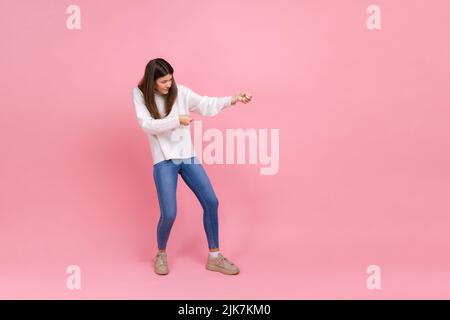 Portrait pleine longueur de fille tirant la corde invisible avec l'effort, en utilisant toute la force pour atteindre le but, en portant blanc style décontracté pull. Studio d'intérieur isolé sur fond rose. Banque D'Images