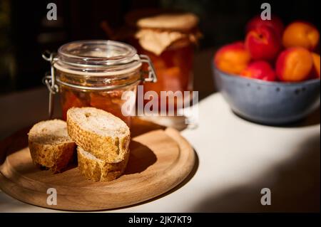 Encore la vie. Tranches de pain à grains entiers sur une planche en bois, un pot avec confiture maison et abricots mûrs sur une table blanche Banque D'Images