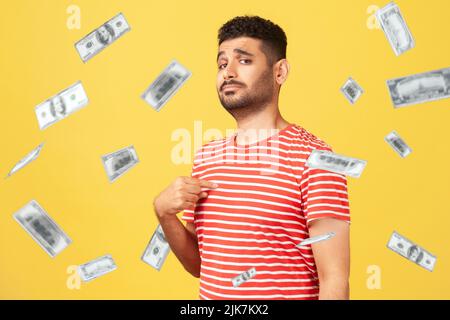 C'est un homme riche. Portrait d'un homme arrogant et confiant, pointant vers la poitrine, regardant fier et égoïste. Argent tombant et il est riche. Studio en intérieur tourné isolé sur fond jaune Banque D'Images