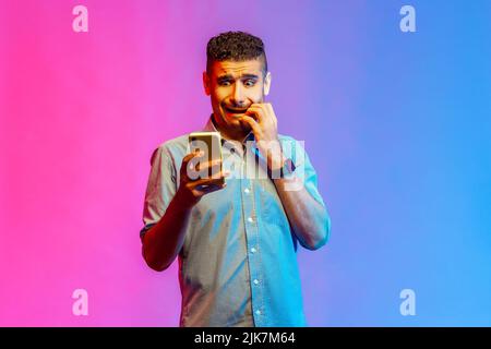 Homme effrayé dans une chemise tenant le téléphone mobile, regardant l'affichage avec peur, piquant ses ongles de doigt, voit quelque chose de terrible. Studio d'intérieur isolé sur fond de néon coloré. Banque D'Images