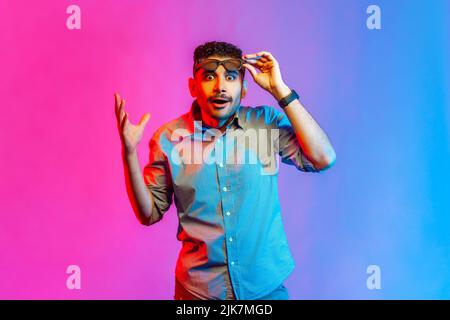 Portrait d'un homme surpris dans une chemise en hauteur lunettes et main, regardant la caméra avec les grands yeux et l'expression choquée. Studio d'intérieur isolé sur fond de néon coloré. Banque D'Images