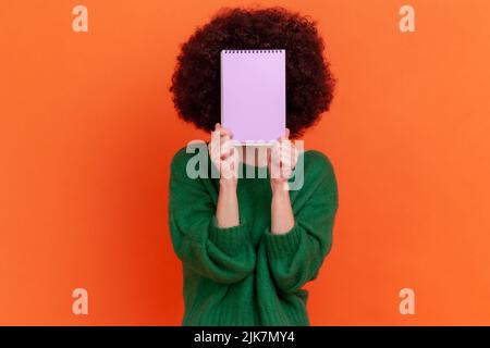 Portrait d'une femme inconnue avec une coiffure afro portant un chandail vert décontracté tenant un organisateur, cachant son visage derrière un carnet en papier. Studio d'intérieur isolé sur fond orange. Banque D'Images