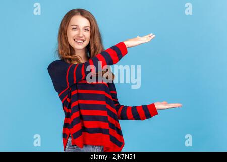 Femme optimiste portant un chandail rayé de style décontracté, présentant l'espace de copie entre les mains, tenant un endroit vide pour le texte ou l'image commerciale. Studio d'intérieur isolé sur fond bleu. Banque D'Images