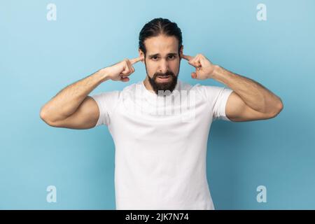 Je ne veux pas ici. Portrait d'un homme avec une barbe portant un T-shirt blanc debout et tenant les doigts sur son oreille, sons désagréables. Studio d'intérieur isolé sur fond bleu. Banque D'Images