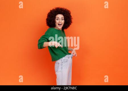 Bonne femme avec la coiffure afro portant vert décontracté style chandail montrant la taille mince dans le grand pantalon, perte de poids réussie, conception de régime. Studio d'intérieur isolé sur fond orange. Banque D'Images