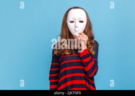 Anonyme femme inconnue portant un chandail rayé style décontracté, debout cache face derrière le masque blanc, complot et intimité, secrets. Studio d'intérieur isolé sur fond bleu. Banque D'Images