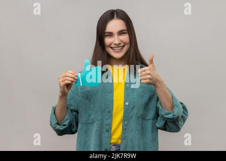 Femme blogueuse tenant le papier bleu pouces vers le haut signe, demandant à évaluer ses postes dans les réseaux sociaux, heureux expression, portant une veste de style décontracté. Prise de vue en studio isolée sur fond gris. Banque D'Images