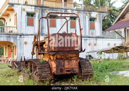 Un ancien tracteur à chenilles rouillé se trouve dans une pelouse surcultivée devant une intéressante maison thaïlandaise de style chinois construite en 1953 Banque D'Images