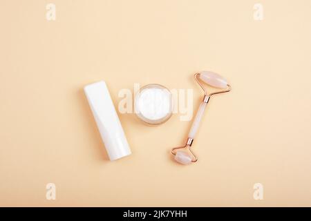 Rouleau facial en cristal de quartz rose, pot crème et tube sur fond beige neutre. Cosmétiques naturels, concept de soins de la peau. Vue de dessus, plan d'installation, maquette. Banque D'Images