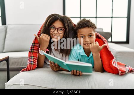 Deux frères et sœurs allongé sur le canapé lisant un livre pointant vers l'arrière avec la main et les pouces vers le haut, souriant confiant Banque D'Images