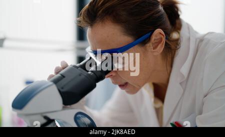 Femme hispanique d'âge moyen portant l'uniforme scientifique au microscope en laboratoire Banque D'Images