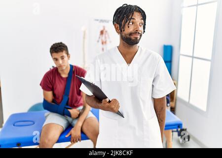 Jeune homme hispanique travaillant à la clinique de récupération de la douleur avec un homme avec un bras brisé souriant regardant le côté et regardant loin de penser. Banque D'Images