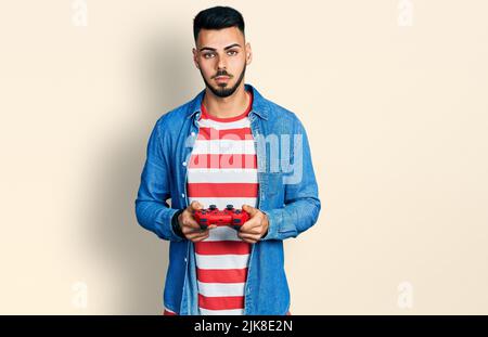 Jeune homme hispanique avec la barbe jouant le jeu vidéo tenant la manette détendue avec l'expression sérieuse sur le visage. Simple et naturel regardant la caméra. Banque D'Images